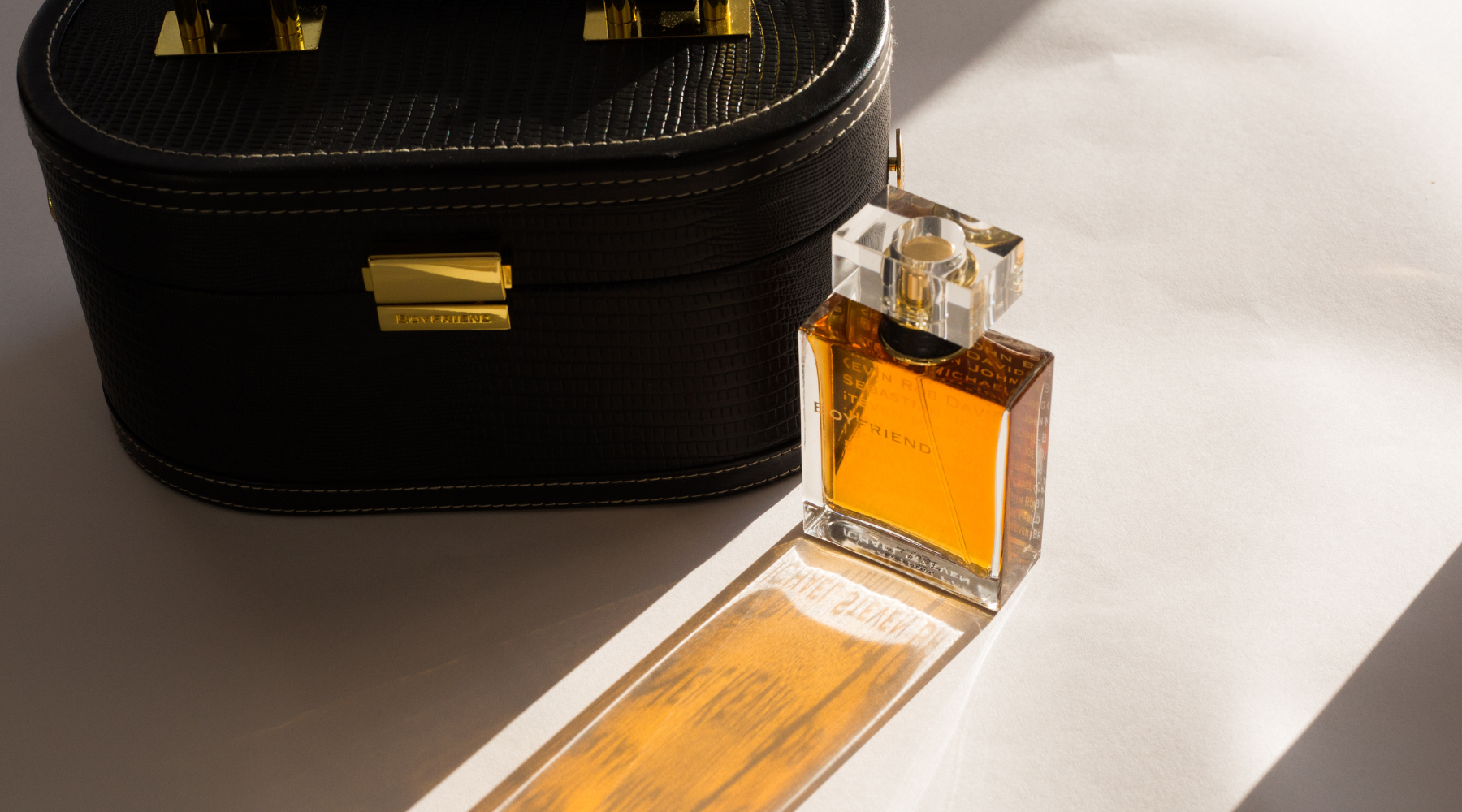 Boyfriend perfume bottle with amber fragrance beside a black leather train case, sunlit.