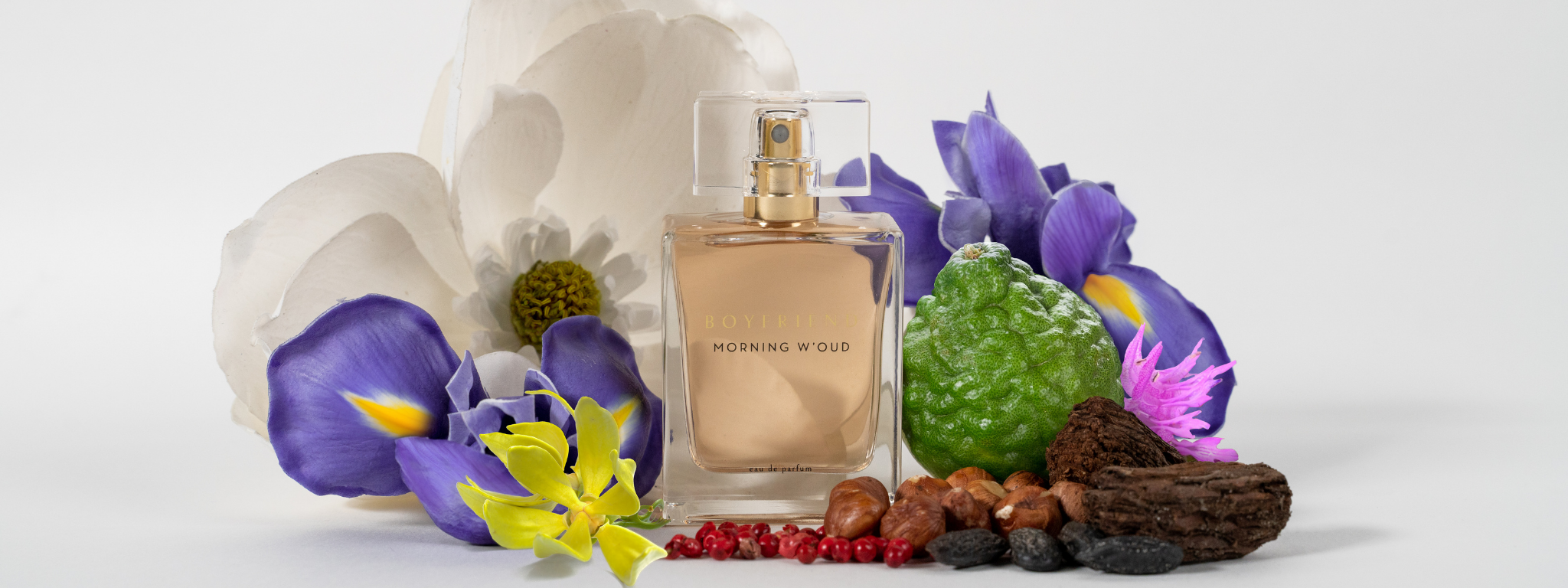 Boyfriend Morning W’oud Eau de Parfum bottle surrounded by magnolia and iris flowers, bergamot fruit, hazelnuts, pink peppercorns, and oud wood against a soft neutral background.